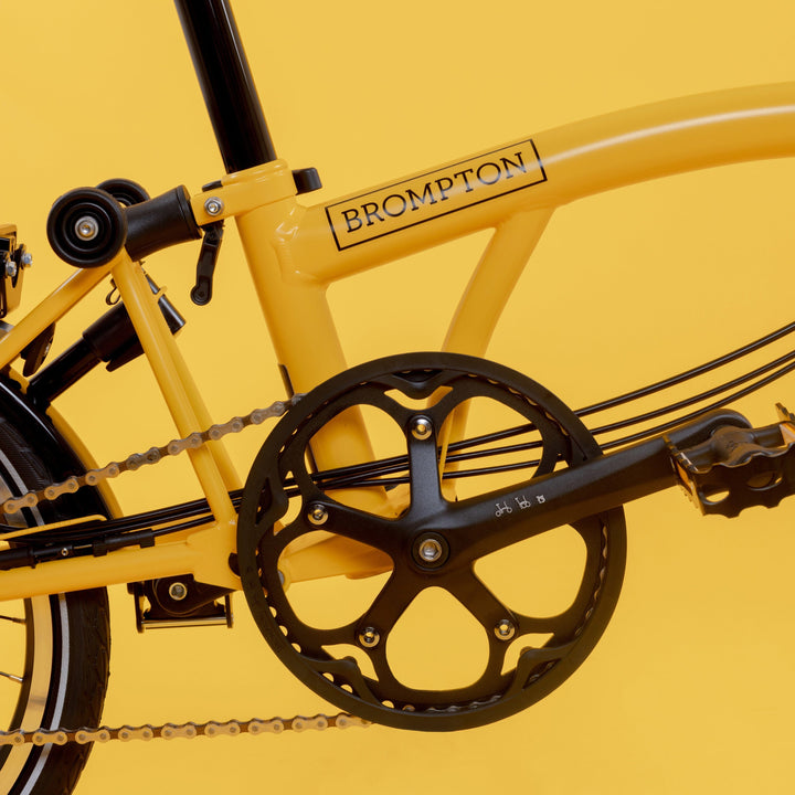 Crankset and Lower Frame of Brompton British Folding Bike in Bright Bumblebee Yellow Colour with a Mid Handlebar on Yellow Background